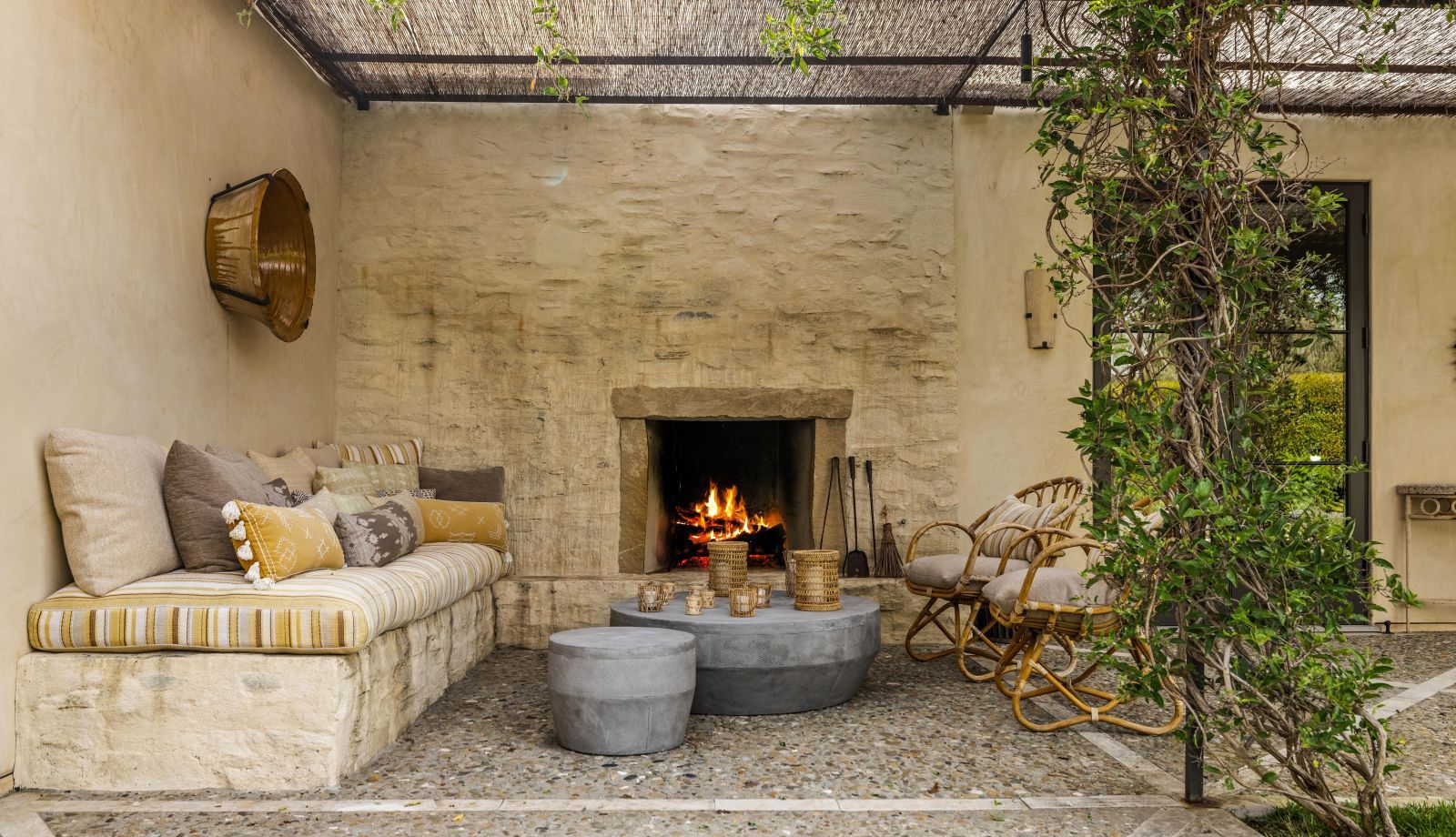 A banquette with a rustic fireplace, rattan chairs, and climbing plant under a pergola